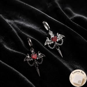 Pode incluir: Um par de brincos de argola com uma pedra vermelha em forma de coração ladeada por asas de morcego prateadas, acentuada com pequenas correntes, uma pedra preta em forma de gota e um pingente pontiagudo. Uma etiqueta diz Custom Box.