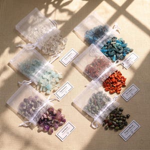 May include: Six white organza drawstring bags filled with tumbled gemstones, including clear quartz, apatite, aquamarine, red jasper, amethyst, and dragon blood stone. Each bag has a small white rectangular label identifying the stone type.