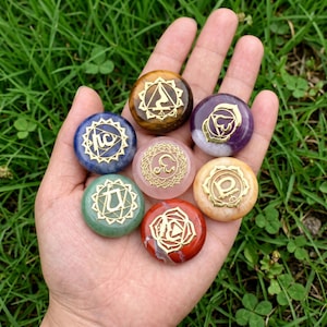 May include: Seven round, polished gemstones in various colors, including tiger's eye, amethyst, rose quartz, sodalite, green aventurine, red jasper, and yellow jade, each featuring a distinct gold-colored metal chakra symbol inlay on the surface.