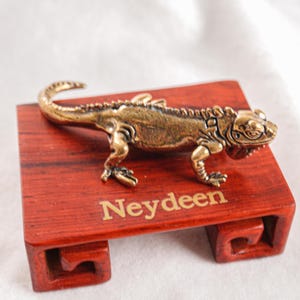 May include: A metallic bronze-toned lizard figurine with textured scales and a curved tail, positioned on a rectangular reddish-brown wooden display stand featuring the word Neydeen printed in gold lettering on the front surface.