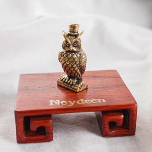 May include: A small brass owl figurine wearing a top hat, perched on a cylindrical branch. The owl is mounted on a rectangular, reddish-brown wooden display stand with carved scroll-like legs and the word Neydeen printed on the top surface.