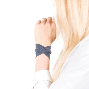 May include: A slate blue wrist cuff with a knotted design. The cuff is made of soft fabric and is worn on the wrist. The background is white.