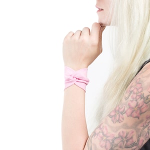 May include: A pink fabric wristband tied in a knot. The wristband is worn on a person's wrist.