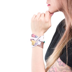 May include: A white wristband with a colorful pattern of cartoon characters and stars. The wristband is tied in a knot and worn on the left wrist.