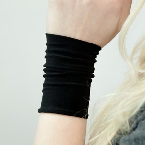 May include: A black wrist cuff made of a soft, stretchy fabric. The cuff is folded over several times to create a layered look.