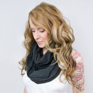 May include: A woman with long, wavy, blonde hair is wearing a dark gray infinity scarf. She is wearing a white shirt and has a floral tattoo on her upper arm.