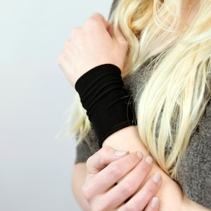 May include: A black wrist cuff made of a soft, stretchy fabric. The cuff is worn on the left wrist of a person with long blonde hair.