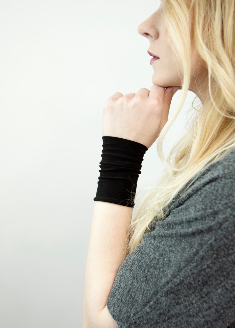 May include: A black wrist cuff made of a soft, stretchy fabric. The cuff is worn on the left wrist of a person with long blonde hair.