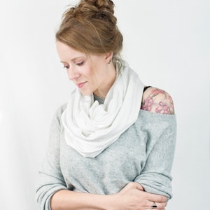 May include: A woman wearing a gray sweater with a white scarf. She has a floral tattoo on her shoulder.
