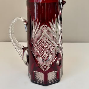 Pode incluir: Um jarro de vidro vermelho rubi com uma alça clara e ornamentada. O jarro apresenta um padrão de diamante e estrela, com um bico para servir. O vidro é cortado com desenhos intrincados, adicionando à sua aparência elegante.