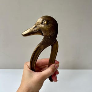 May include: A brass nutcracker shaped like a duck head with a hinged beak mechanism. The metal has a warm, golden-brown patina with detailed etched lines representing feathers on the head and neck, and a smooth finish on the handle sections.