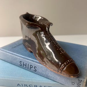 May include: A small, glossy brown ceramic shoe ornament with a lace-up design. The miniature shoe is placed on two light blue books. The word "SHIPS" is visible on the spine of one book.