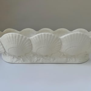 May include: Off-white ceramic seashell-shaped planter. The rectangular planter features a scalloped rim and is decorated with multiple seashell designs. The base of the planter has a textured, wave-like design. The planter is approximately 12 inches long.