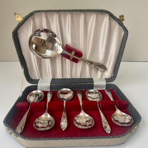 Puede incluir: Un juego de cucharas plateadas vintage, que comprende una cuchara de servir y seis cucharas más pequeñas, presentado en una caja decorativa. Las cucharas tienen mangos detallados y un interior de terciopelo rojo. La caja es gris con un forro de satén blanco.