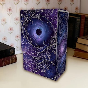 Könnte beinhalten: Eine dekorative Buchhülle mit einem kosmischen Design. Sie zeigt eine wirbelnde Galaxie und Halbmonde in Lila- und Blautönen, umrahmt von silberfarbenen Ranken und Blättern. Die Buchhülle steht auf einer weißen Oberfläche.