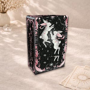 Può includere: Scatola nera per carte dei tarocchi con conigli bianchi con orecchie e dettagli rosa, su sfondo stellato. La scatola ha un bordo floreale decorativo rosa e nero. Carte dei tarocchi visibili.