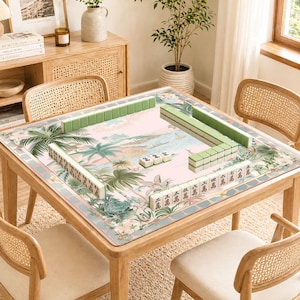 May include: A wooden mahjong table with a tropical landscape design on the playing surface. Green and white mahjong tiles are arranged on the table. The table is surrounded by four rattan chairs.