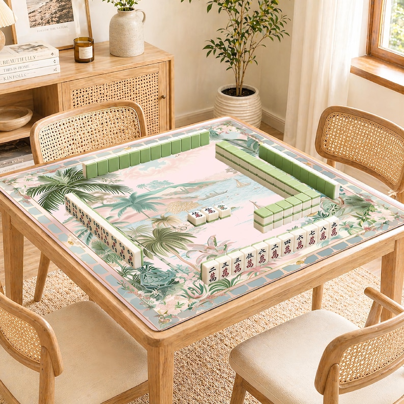 May include: A wooden mahjong table with a tropical landscape design on the playing surface. Green and white mahjong tiles are arranged on the table. The table is surrounded by four rattan chairs.