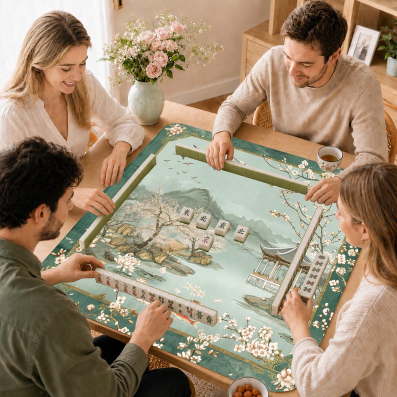May include: A square mahjong mat featuring a light green landscape illustration with cherry blossoms, a traditional pavilion, and mountains. The mat has a dark green border with floral patterns and holds several mahjong tiles with Chinese characters.