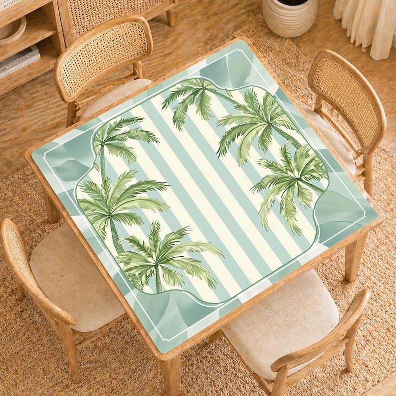 Pode incluir: Uma mesa quadrada de madeira com um design tropical. O tampo da mesa apresenta um fundo listrado azul claro e branco com uma borda de palmeiras verdes. Quatro cadeiras de madeira com encostos tecidos circundam a mesa.