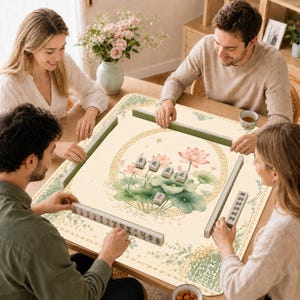 May include: A square mahjong mat featuring a cream-colored surface with a central illustration of pink lotus flowers and green lily pads enclosed in a circular gold border. The mat includes green tile racks and white mahjong tiles with black and red characters.