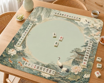Puede incluir: Una alfombrilla de mahjong cuadrada con un círculo central verde claro rodeado por una ilustración paisajística detallada de peces koi, flores de loto, grullas y un pabellón. La alfombrilla está dispuesta con fichas de mahjong blancas en los bordes.