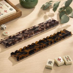 Puede incluir: Dos soportes rectangulares para fichas de mahjong con un patrón de carey marrón y negro. Un soporte presenta una superficie lisa e inclinada, mientras que el otro tiene un diseño de borde festoneado en la parte frontal para sujetar las fichas.