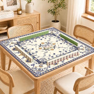 May include: A square wooden table with a cream-colored Mahjong game mat featuring a blue floral design. Green and white Mahjong tiles are arranged around the mat. The table is surrounded by four rattan chairs.