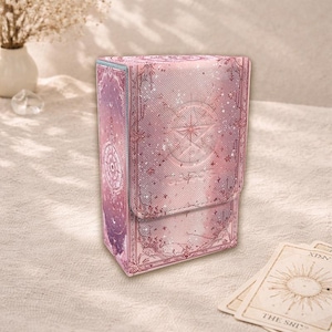 Op de afbeelding: Een roze tarotkaartendoos met een hemels ontwerp. De doos heeft een sterrenkompas en bloemaccenten. De doos is rechthoekig met een magnetische klepsluiting. Tarotkaarten zijn zichtbaar op de voorgrond.