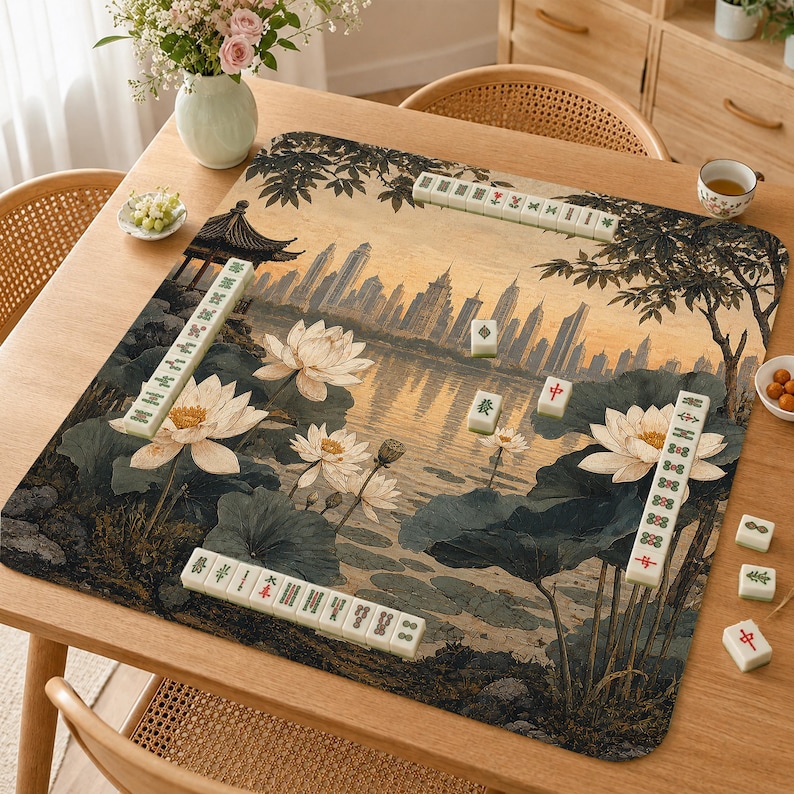 May include: A square mahjong mat featuring a printed scene of a lotus pond with large white flowers and green leaves in the foreground, set against a city skyline at sunset. Several rows and individual white mahjong tiles are placed on the mat.