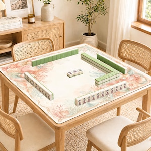 May include: A wooden square table with a floral patterned mat, set for a game of Mahjong. Green and white tiles are arranged on the mat. The table is surrounded by rattan chairs, with a wooden cabinet and decor in the background.