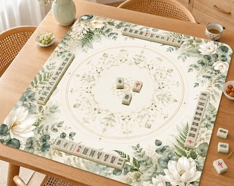 May include: A square mahjong mat featuring a cream-colored center with a delicate gold circular mandala design. The border is decorated with a botanical print of white lotus flowers, green eucalyptus leaves, and ferns. Several mahjong tiles are arranged.