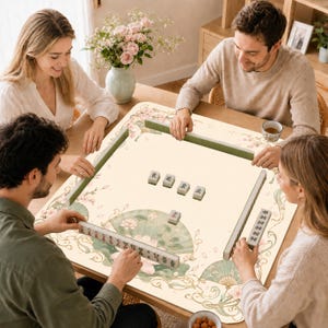 Puede incluir: Un tapete de mahjong cuadrado con una superficie de color crema, decorado con delicados patrones florales, ilustraciones de abanicos verdes y bordes decorativos. El tapete muestra varias fichas de mahjong dispuestas en filas y dispersas.