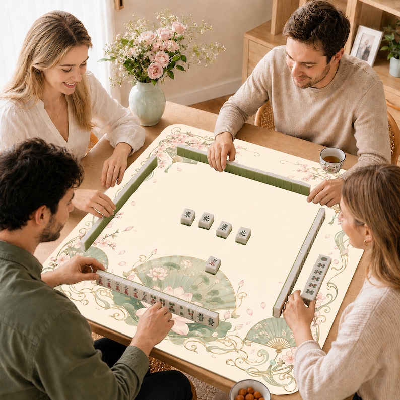 Puede incluir: Un tapete de mahjong cuadrado con una superficie de color crema, decorado con delicados patrones florales, ilustraciones de abanicos verdes y bordes decorativos. El tapete muestra varias fichas de mahjong dispuestas en filas y dispersas.