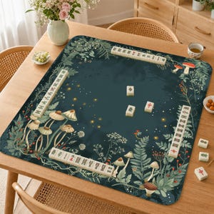 Pode incluir: Um tapete de mahjong quadrado em verde-azulado escuro com uma estampa botânica de cogumelos, fetos e folhagens com pirilampos espalhados. O tapete exibe quatro filas de peças de mahjong em marfim nas bordas e três peças soltas no centro.