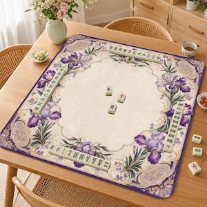 Puede incluir: Un tapete cuadrado de mahjong con un centro color crema y un borde decorativo de flores de iris moradas y volutas doradas. El tapete está dispuesto con filas de fichas de mahjong color marfil a lo largo de los bordes y tres fichas en el centro.