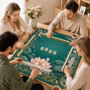 Op de afbeelding: Een vierkante mahjongmat met een diepe blauwgroene achtergrond, versierd met een grote roze lotusbloem, groene waterleliebladeren en gouden pauwenveerpatronen. De mat toont vier witte mahjongstenen in het midden en rekjes met stenen.
