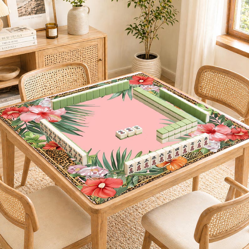 May include: A wooden mahjong table with a pink playing surface, edged with floral and leopard print borders. Green and white mahjong tiles are arranged on the table. The table is surrounded by four rattan chairs. The table is approximately 100 cm square.