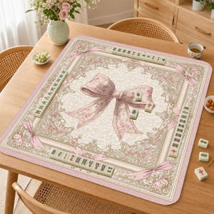Könnte beinhalten: Ein rosa-cremefarbener Mahjong-Spielteppich mit einem großen Schleifendesign in der Mitte. Der Teppich ist von einem floralen Rand und rosa Bändern umgeben. Mehrere Mahjong-Steine sind um den Teppich herum platziert, spielbereit.