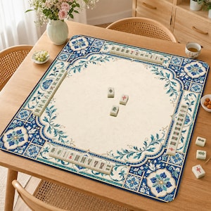 May include: A square mahjong mat featuring a cream-colored center and a decorative border with blue and white floral tile patterns. The mat displays rows of ivory-colored mahjong tiles along the edges and three individual tiles in the center.