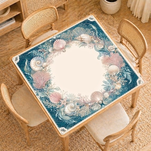 May include: Square wooden table with a marine-themed design. The table features a cream-colored center surrounded by a border of seashells, coral, and pearls in shades of pink, blue, and beige. The table is surrounded by four wooden chairs.