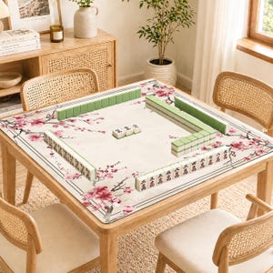 May include: A wooden square mahjong table with a floral patterned mat featuring pink blossoms. Green and white mahjong tiles are arranged on the table. The table is surrounded by four rattan chairs.