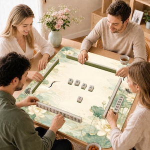 May include: A square mahjong mat featuring a light cream background with a green and white lotus flower and crane pattern. The mat is set on a wooden table with several mahjong tiles arranged in the center and along the edges of the surface.