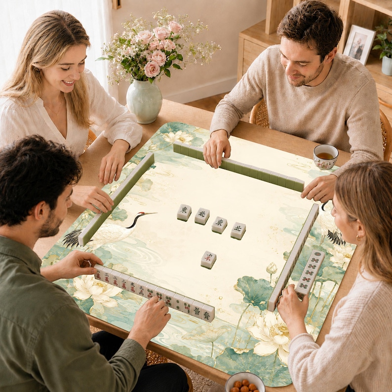May include: A square mahjong mat featuring a light cream background with a green and white lotus flower and crane pattern. The mat is set on a wooden table with several mahjong tiles arranged in the center and along the edges of the surface.