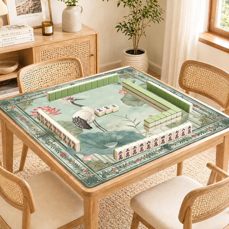 May include: A wooden square Mahjong table with a green and floral patterned mat. The mat features a crane, lotus flowers, and Chinese characters. Green and white Mahjong tiles are arranged on the table, surrounded by four rattan chairs.