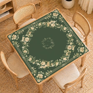 May include: A square, dark green table cover with a floral design. The border features a cream-colored geometric pattern and a garland of cream-colored roses and foliage. The table is surrounded by four wooden chairs with woven backs.