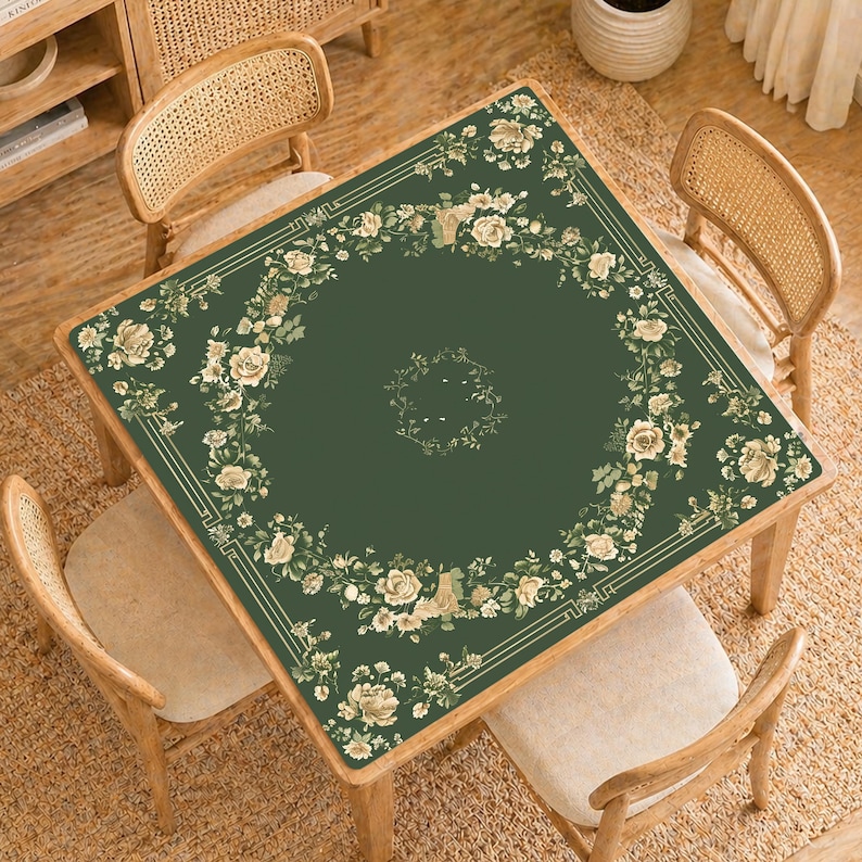 May include: A square, dark green table cover with a floral design. The border features a cream-colored geometric pattern and a garland of cream-colored roses and foliage. The table is surrounded by four wooden chairs with woven backs.