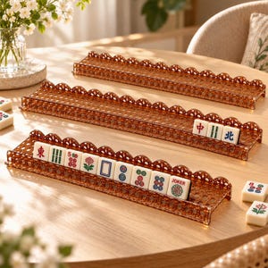 May include: Three brown translucent plastic Mahjong tile racks featuring a decorative scalloped edge. The racks are arranged on a wooden surface, with several white Mahjong tiles displayed on the front and middle racks, showing various symbols and the word JOKER.