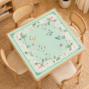 Puede incluir: Una mesa cuadrada con un mantel verde claro con un diseño floral y de mariposas. El mantel tiene un borde festoneado y un borde a cuadros. Rosas rosas y mariposas están esparcidas por la superficie. Cuatro sillas de madera rodean la mesa.