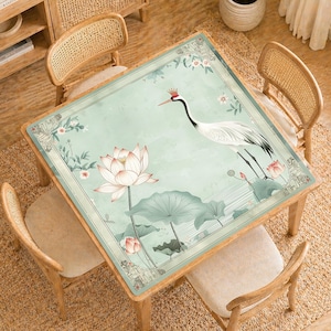 May include: Square wooden table with a light green floral design featuring a crane, lotus flowers, and leaves. The table is surrounded by four wooden chairs with woven backs and beige cushions. The table is set on a woven rug.